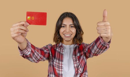 A young middle eastern woman is smiling and giving a thumbs up while holding a red credit card in front of her. She is wearing a plaid shirt and is standing against a light brown background.