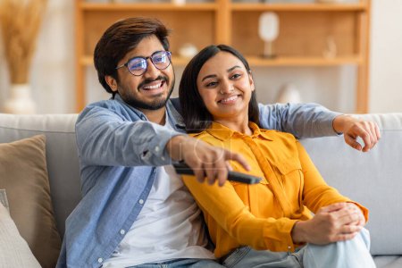 A smiling Indian couple is sitting on a sofa, enjoying their afternoon while watching television. They appear relaxed and happy, with the man holding a remote control