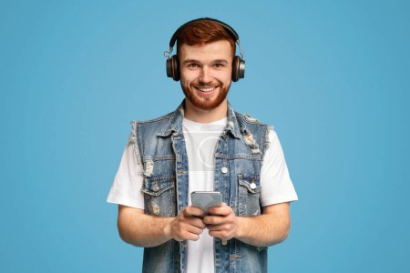 Partager une super chanson avec des amis. Jeune homme roux souriant écoutant de la musique et tenant son téléphone portable sur fond bleu, espace de copie