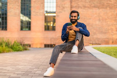 L'Indien est assis sur un banc dans un parc urbain, profitant du beau temps. Il porte des écouteurs et regarde attentivement son smartphone, souriant.