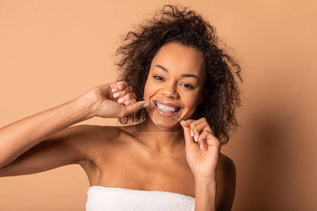 Eine junge afroamerikanische Frau mit lockigem braunem Haar lächelt, als sie mit Zahnseide zwischen ihren Zähnen putzt. Sie trägt ein weißes Handtuch und steht vor braunem Hintergrund