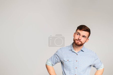 Nachdenklicher junger bärtiger Mann in lässigem Hemd, der den Kopierraum auf grauem Studiohintergrund betrachtet