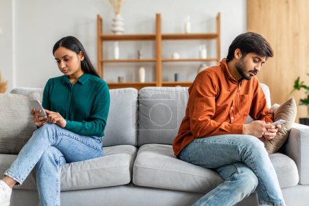 Indian couple sits on a gray couch in a modern living room, both absorbed in their smartphones. The room features light-colored walls, wooden shelves, and minimalistic decor