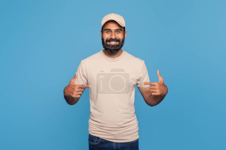 A happy bearded Indian man in a beige shirt and matching cap stands against a blue background, smiling and pointing at his t-shirt, conveying a sense of pride and joy.