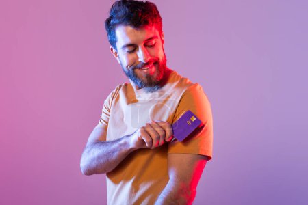 A cheerful man with a beard holds his credit card and smiles, wearing an orange shirt. The background features vibrant pink and purple hues.