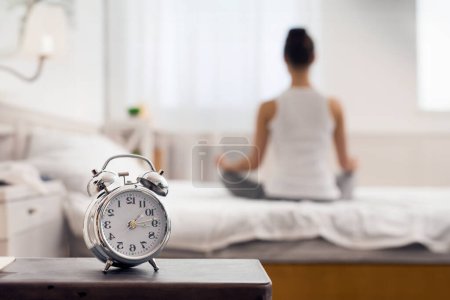 Morning yoga. Alarm clock standing on bedside table, woman meditating on bed
