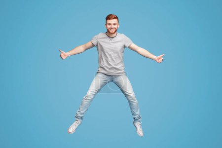 Young millennial redhaired guy jumping in air on blue background, demonstrating thumbs up gesture, full length, studio, empty space