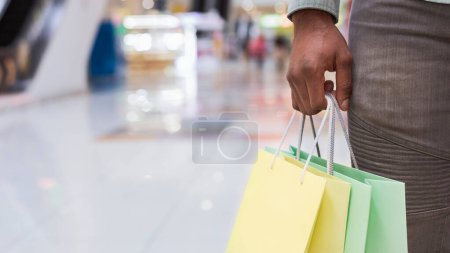 Cultivado de hombre afroamericano con ropa casual se ve de pie en un bullicioso centro comercial, sosteniendo múltiples bolsas de compras en sus manos, rodeado de tiendas y otros compradores.