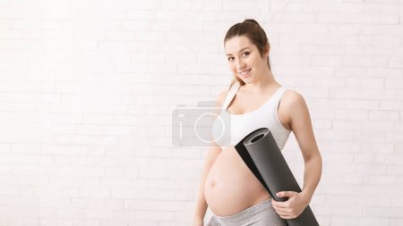 Sportliche Schwangere steht da und hält eine Yogamatte in den Händen. Sie ist sichtlich erwartungsvoll und scheint sich auf eine Yoga-Stunde vorzubereiten.
