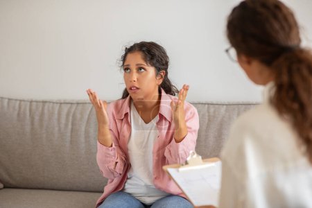 A young woman sits on a couch during therapy session, expressing her frustration with her hands up in the air, appears stressed and agitated, while the therapist listens intently, holding clipboard.
