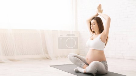 A pregnant woman sits cross-legged on a yoga mat, practicing a meditation pose with her hands together over her head. The bright, airy living room enhances the serene morning atmosphere