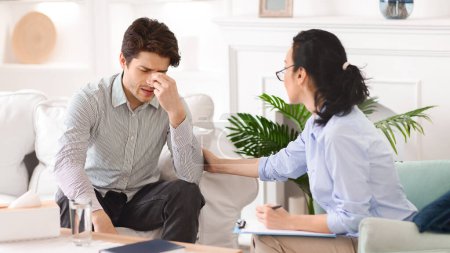 A man sits on a white couch, visibly distressed, holding his forehead while a therapist provides support. The session occurs in a calmly decorated office with modern furnishings and greenery.