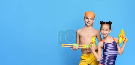 Two cheerful children, dressed in bright swimwear and headbands, are smiling while holding colorful water guns, ready for some fun activities, panorama with copy space