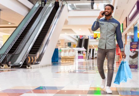A black man is seen walking through a bustling shopping mall, engaged in a phone conversation. He appears focused on his call amidst the hustle and bustle of the malls activity, copy space