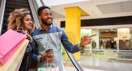 African American man and woman are seen browsing through various stores in a shopping mall, holding shopping bags and looking at products, panorama with copy space