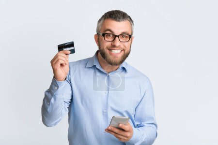 A man is depicted holding a credit card in one hand and a cell phone in the other hand. He appears to be engaged in a transaction or making a purchase online.