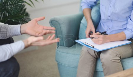 Cultivado de terapeuta escucha activamente mientras toma notas en un portapapeles, durante una sesión de asesoramiento con el hombre en una oficina moderna en una tarde soleada