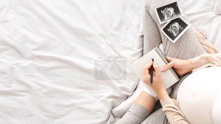 A pregnant woman is seated comfortably in bed, writing in a journal. Beside her are ultrasound images of her unborn baby, top view, copy space