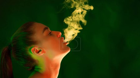 A woman is standing in front of a plain green background, smoking a cigarette. She is blowing smoke into the air, copy space