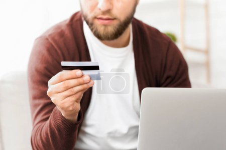 A man is shown holding a credit card in one hand and a laptop in the other. He appears to be in the process of making an online purchase or managing his finances, cropped