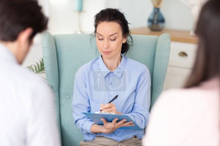 A woman counselor attentively listens to two clients spouses and takes notes during a morning therapy session in a well-lit, comfortable clinic room.