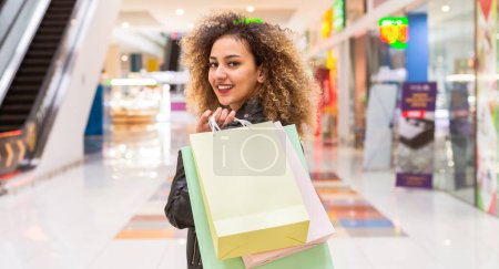 Eine junge Afroamerikanerin ist mit mehreren Einkaufstüten in einem belebten Einkaufszentrum zu sehen. Sie sieht konzentriert aus, als sie durch die geschäftige Umgebung navigiert, umgeben von verschiedenen Geschäften..