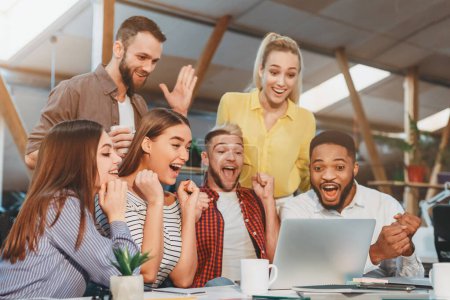 A diverse group of young professionals cheer and celebrate after achieving a milestone while gathered around a laptop in a contemporary office setting.