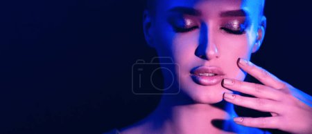A woman closes her eyes and touches her face gently, bathed in dramatic blue and purple lighting, creating a contemplative mood, panorama with copy space