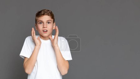 Un adolescente con una expresión impactada sostiene sus mejillas aisladas sobre un fondo gris, mostrando una gama de emociones de los jóvenes, espacio de copia