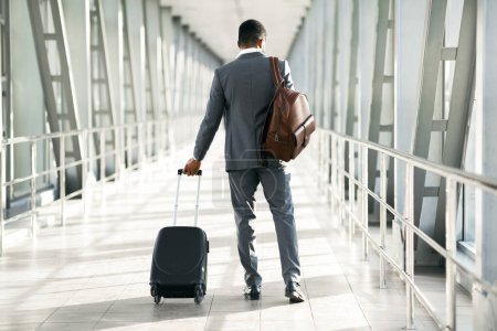 Un viajero corporativo con equipaje es visto en un aeropuerto. El estado de ánimo es enfocado y profesional, con un entorno moderno en el aeropuerto que enfatiza los viajes y negocios, vista trasera