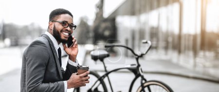 Smiling African American businessman holding coffee cup and phone, sitting near a bicycle in urban setting, web-banner