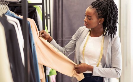 Mujer afroamericana de pie delante de un estante de ropa, examinando cuidadosamente las diferentes prendas que se muestran. Ella sostiene algunos artículos en sus manos mientras navega a través de la selección.