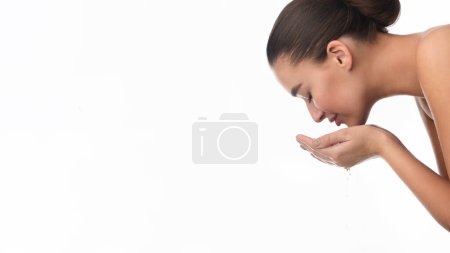Eine Frau steht vor einem Waschbecken und spritzt sich mit den Händen Wasser ins Gesicht. Sie trägt ein weißes T-Shirt und hat die Haare nach hinten gezogen. Die Wassertropfen sind auf ihrer Haut zu sehen.