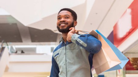 El hombre negro positivo es visto sosteniendo múltiples bolsas de compras mientras camina a través de un bullicioso centro comercial. Parece concentrado en sus compras, maniobrando a través de la multitud con facilidad.