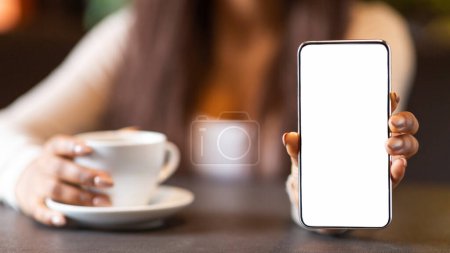 A girl is enjoying a cup of coffee in a cozy cafe while holding a white smartphone with a blank screen towards the camera, cropped, mockup, copy space