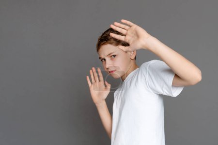 In this image, a teen boy adopts a serious expression and makes a stop gesture with his hands, signaling a boundary or refusal, isolated on a grey background