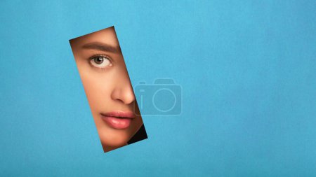 Beautiful woman face is visible through a hole in a blue wall, looking out with curiosity or surprise. The opening provides a glimpse into the enclosed space, copy space