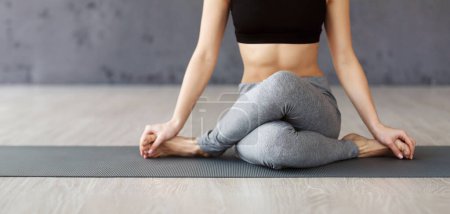 A woman is seated in a cross-legged position on a yoga mat, performing a yoga pose in a bright studio during the morning.