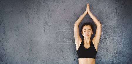 Eine junge Frau steht vor einer grauen Wand, die Hände über dem Kopf zusammengepresst, die Augen geschlossen, und praktiziert frühmorgens Yoga-Meditation.