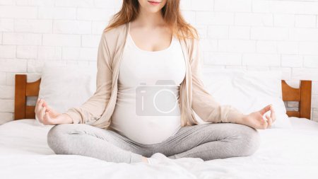 A pregnant woman is sitting cross-legged on a bed, in deep concentration as she practices meditation, creating a serene and focused atmosphere in the room, cropped