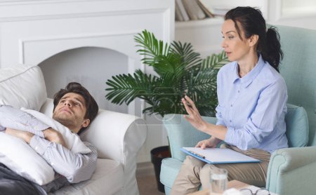 A therapist engages with a man patient lying on a sofa, providing counseling in a contemporary living room with natural light and indoor plants.