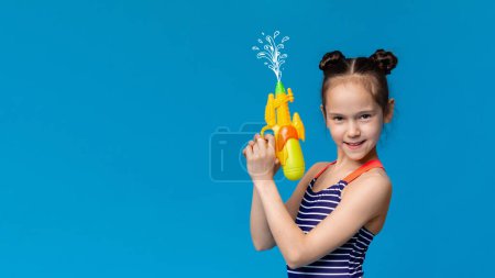 Mädchen in Badebekleidung schießt mit Wasserpistole hoch, blauer Studiohintergrund