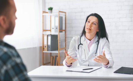 Woman doctor in a white coat and stethoscope is sitting at a desk in an office, explaining medical information to a patient who is listening attentively. The doctor has a serious expression