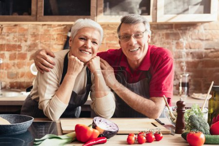 Ein älteres Ehepaar steht nebeneinander in der Küche und bereitet eine Mahlzeit zu. Die Frau schneidet eine Gurke auf einem Schneidebrett, während der Mann eine gelbe Paprika hochhält. Beide tragen Schürzen