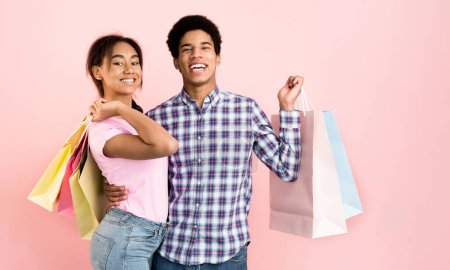 Ein junger schwarzer Mann und ein junges Mädchen stehen vor rosa Hintergrund und halten jeweils Einkaufstüten in der Hand. Sie scheinen auf einer Einkaufstour gewesen zu sein, wobei die Taschen darauf hindeuten, dass sie möglicherweise eingekauft haben..
