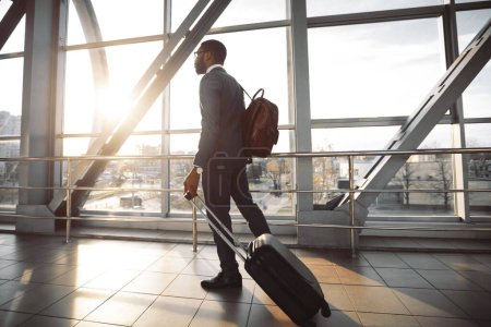 El hombre de negocios negro está tirando de su maleta en el exterior de un aeropuerto moderno. La escena es animada y profesional, reflejando la eficiencia y el ajetreo de los viajes de negocios..