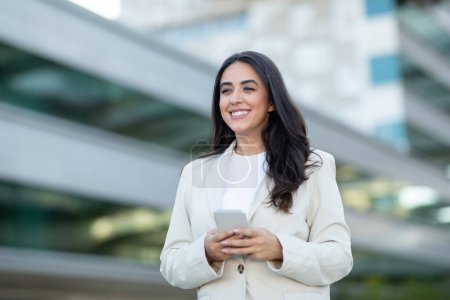 A smiling woman with long dark hair is dressed in a white blazer and a white t-shirt. She is standing in an urban setting with a modern office building in the background, holding her smartphone