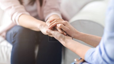 Une femme offre confort et réconfort en tenant une autre main de femme lors d'une conversation sincère dans un cadre confortable salon, recadré