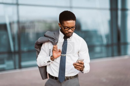 Un hombre de negocios negro viajante está usando su teléfono mientras está parado fuera de un aeropuerto contemporáneo. El estado de ánimo es centrado y eficiente, destacando los viajes y los negocios.