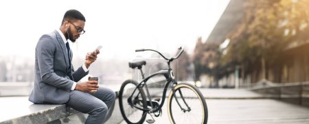 Un hombre de negocios afroamericano está sentado junto a su bicicleta mientras usa su teléfono. El ambiente es relajado y profesional, ambientado en un entorno urbano al aire libre.